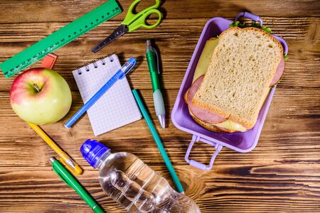 Bottle of water, ripe apple, different stationeries and lunch box with sandwiches on wooden table. Top viewの写真素材