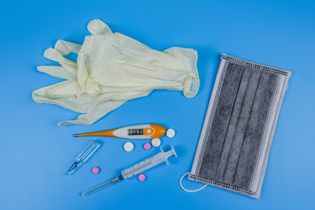 Medical digital thermometer, protective face mask, pills, and syringe on blue background. Healthcare...