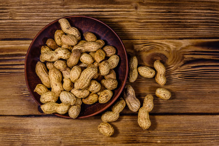 Ceramic plate with heap of the unpeeled peanuts on a wooden table. Top view