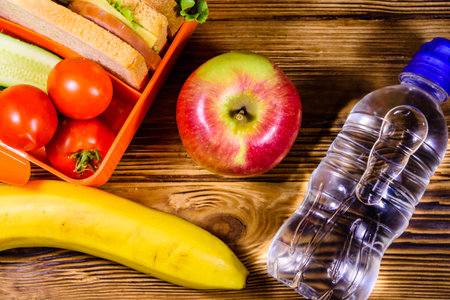 Ripe apple, banana, bottle of water and lunch box with sandwiches, cucumbers and tomatoes on rustic wooden table. top viewの写真素材