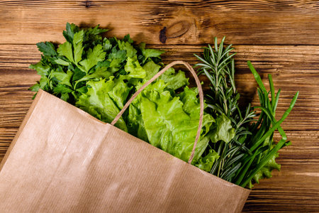 Paper bag with lettuce rosemary and parsley on wooden table. Vegan food. Top viewの写真素材