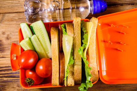 Bottle of water and lunch box with sandwiches, cucumbers and tomatoes on rustic wooden table. Top viewの写真素材