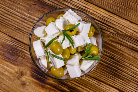 Chopped feta cheese, rosemary and olives in glass bowl on wooden table. Top view