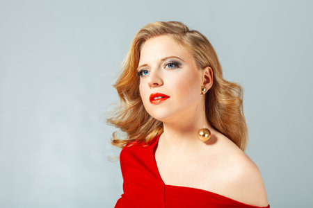A glamorous woman with red lips, styled blond hair, and striking makeup poses in a red dress. Her bold gold earrings and confident look highlight her elegance. Background is neutral, emphasizing her sophisticated style.の写真素材