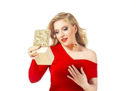 Elegant woman wearing a red dress and gold earrings, holding a decorative gift box. Her blond hair and bold makeup enhance her glamorous appearance. Perfect for holiday and luxury concepts.の写真素材