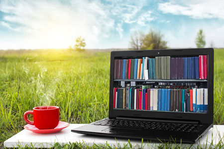 Laptop and cup of hot coffee on the background picturesque nature, outdoor office. E-book library concept with laptop computer and book. Old books on a wooden shelf. Travel concept. Business ideas.の写真素材