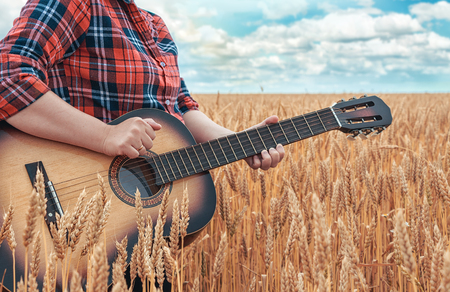 Girl in the red shirt in wheat field plays the acoustic guitar. Beautiful nature at bright sunny summer day. Camping.の写真素材