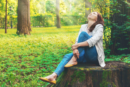 Beautiful girl sits on a big old stump in the forest, the sun's rays breaking through the leaves and illuminate her face. Fabulous country. Cute woman is enjoying of picturesque nature. Camping, rest.の写真素材