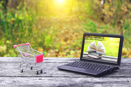 Shopping cart and laptop with books on the screen on the old wooden table at the nature background. Concept: back to school, education. Online purchase.の写真素材