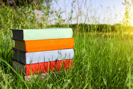 Stack of multicolored books on the green grass on the background of beautiful nature surrounded by meadows at sunny day.Library at outdoor. Back to school.の写真素材
