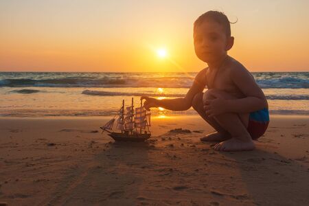 Little baby boy is playing with toy ship on the sand of amazing tropical beach of Andaman sea in Thailand at sunset. Concept of travel with children. Family holidays.の写真素材