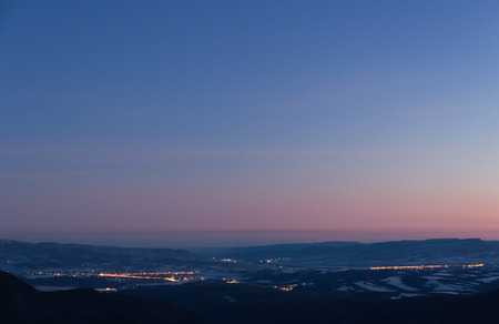 The winter early morning view from the mountains to a valley with towns emitting light and with blue pink sky slightly covered by clouds の写真素材