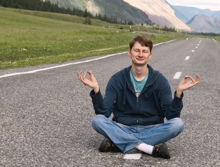 Portrait of an attractive young caucasian man (guy, male, person, model) sitting on the asphalt road among mountain landscape  in the lotus cross-legged position meditating with closed eyes and smiling.の写真素材
