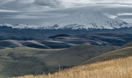 Snow-capped Elbrus mountain surrounded by clouds rises above the mountain area in Kabardino-Balkaria (Caucasus, Russia). Photo has been shot in September, 2014. Can be used as a background, wallpaper or postcard.の写真素材
