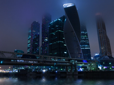 Modern urban architecture: Moscow City commercial center by night in mist and fog illuminated. Includes skyscreper buildings with alight windows and bridge over the Moskva river.の写真素材
