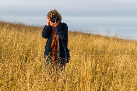 Nice looking pretty blondy caucasian happy successful girl with camera staying within the dry grass landscape shooting somebody.の写真素材