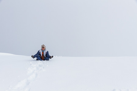 Nice looking pretty blondy caucasian happy successful girl sitting in the zen pose at the top of mountain covered by snow with cloudy sky and mist as a background.の写真素材