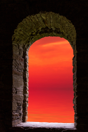 Red sunset evening sky with clouds reflected in wavy water and visible through frame of old ancient stone window.の写真素材