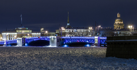 Old architecture landmark and touristic spot in Saint Petersburg, Russia by a winter night: the Neva river covered by ice and snow, the Palace bridge and historical city center illuminated.の写真素材