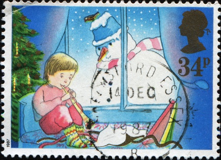 GREAT BRITAIN - CIRCA 1987: A greeting Christmas stamp printed in Great Britain shows boy sitting on the bed under the Christmas tree and playing a pipe, into the window looks a snowman, circa 1987の写真素材