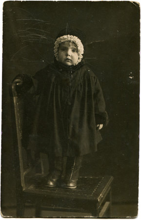 portrait of little girl in a dark coat and a hat with a white frill, standing on a chair, Lugansk,...