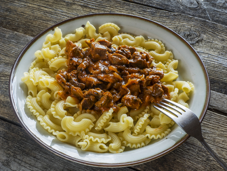 Pasta with meat, stewed in tomato sauce on a plate on old weathered wooden tableの写真素材