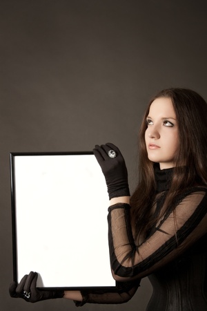 Pretty gothic girl in black dress with blank frame in hands over gray backgroundの写真素材