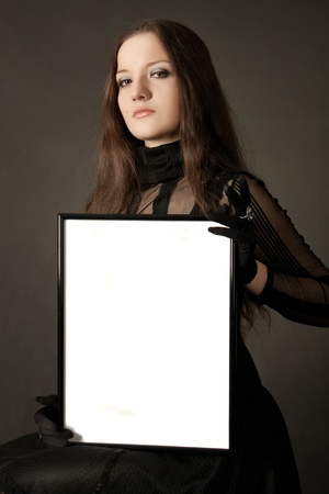 Pretty gothic girl in black dress with blank frame in hands over gray backgroundの写真素材