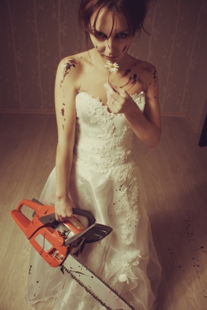 Pretty young bride standing in the empty room with bloody chainsaw and chamomileの写真素材