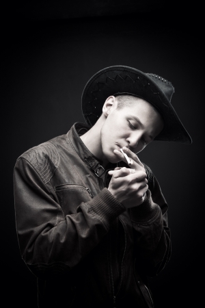 Young man  in hat posing over dark background with cigarette lighter の写真素材