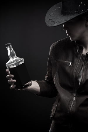 Brutal macho in hat holding bottle of wisky over dark backgroundの写真素材
