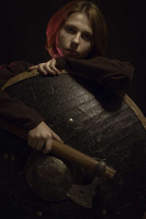 Cute young scandinavian medievail woman posing with shield and axe over black background
