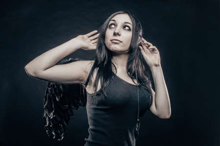 Pretty brunette girl with wings and with headphones posing over dark background.の写真素材