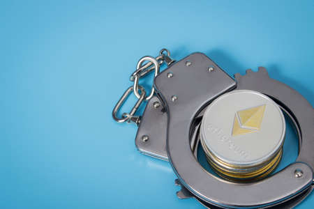 One ethereum coin and handcuffs over blue background.