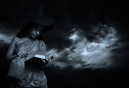 Pretty girl in witch hat posing with book over dramatic skyの写真素材