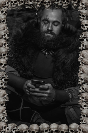 Brutal scandinavian warrior sitting on rocky sea shore with smart phone and headphonesの写真素材