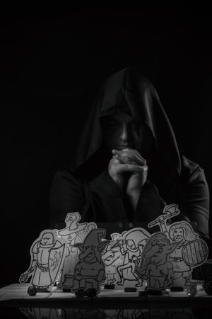 Dark and atmospheric portrait of a man wearing a hooded cloak sitting at a tabletop role-playing...