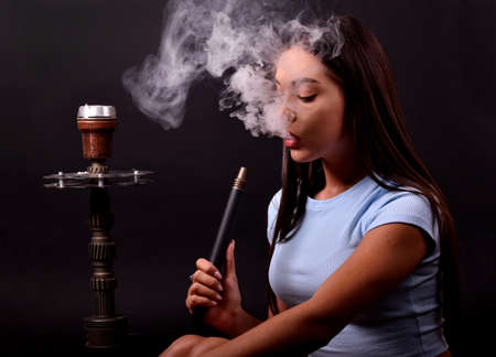 The girl smokes a hookah on a black background. Smoke comes out of the mouth. The pleasure of...