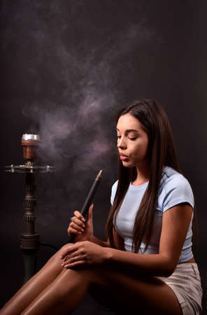 The girl smokes a hookah on a black background. Smoke comes out of the mouth. The pleasure of smokingの写真素材