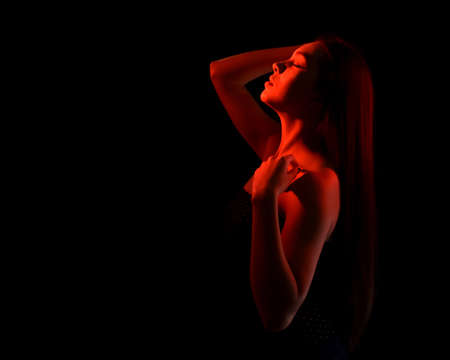 portrait of a beautiful young woman with closed eyes and bare shoulders, isolated on black background with red light. space for textの写真素材
