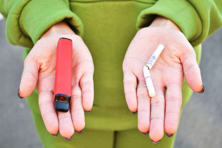 Close-up view of female hands holding an e-cigarette in one hand and nicotine cigarette in another one. Choice concept.Differences in taste and preferenceの写真素材