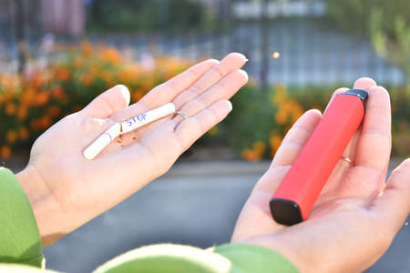Close-up view of female hands holding an e-cigarette in one hand and nicotine cigarette in another one. Choice concept.Differences in taste and preferenceの写真素材