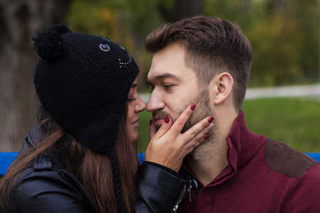 Closeup shot of young beautiful stylish couple in autumn park. She is kissing him. He has a beard. She has a piercing in the nose. She wears black hat with bubo.の写真素材