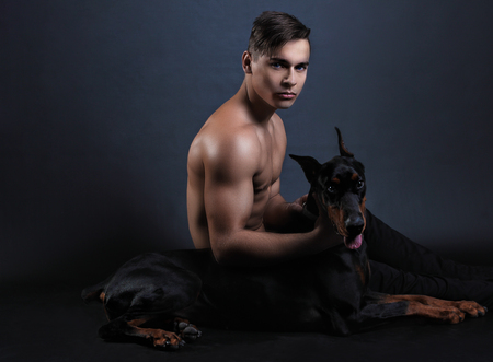muscular man with a naked torso sitting with black doberman dog on black backgroundの写真素材