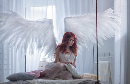beautiful young model with open angel wings and pink dress sitting on hanging bed suspended from the ceiling. White loft interior.の写真素材