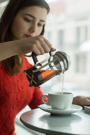  Young girl pours black tea from glass kettle to smal cup in cafe with big windowの写真素材