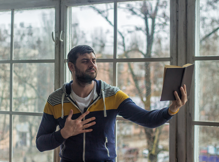 Bearded poet man reads a book with an expression. Big window on background.