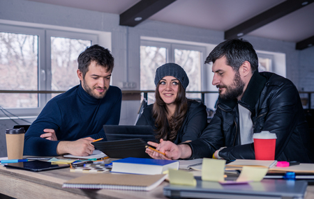 Young smart team. Group of  modern people discussing work on tablet in the office. Working environment with laptop, coffee, notepads and stationery. They are looking at the screen. Business team concept.の写真素材