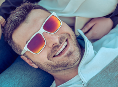 Smiling happy bearded man wearing Colorful sunglasses lying on his girlfriend. He is looking at the cameraの写真素材
