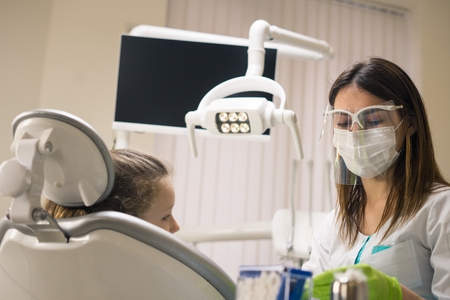 Dentist woman preparing  for treatment  at dental clinic. She is wearing dental facemask. She has little girl patient.の写真素材